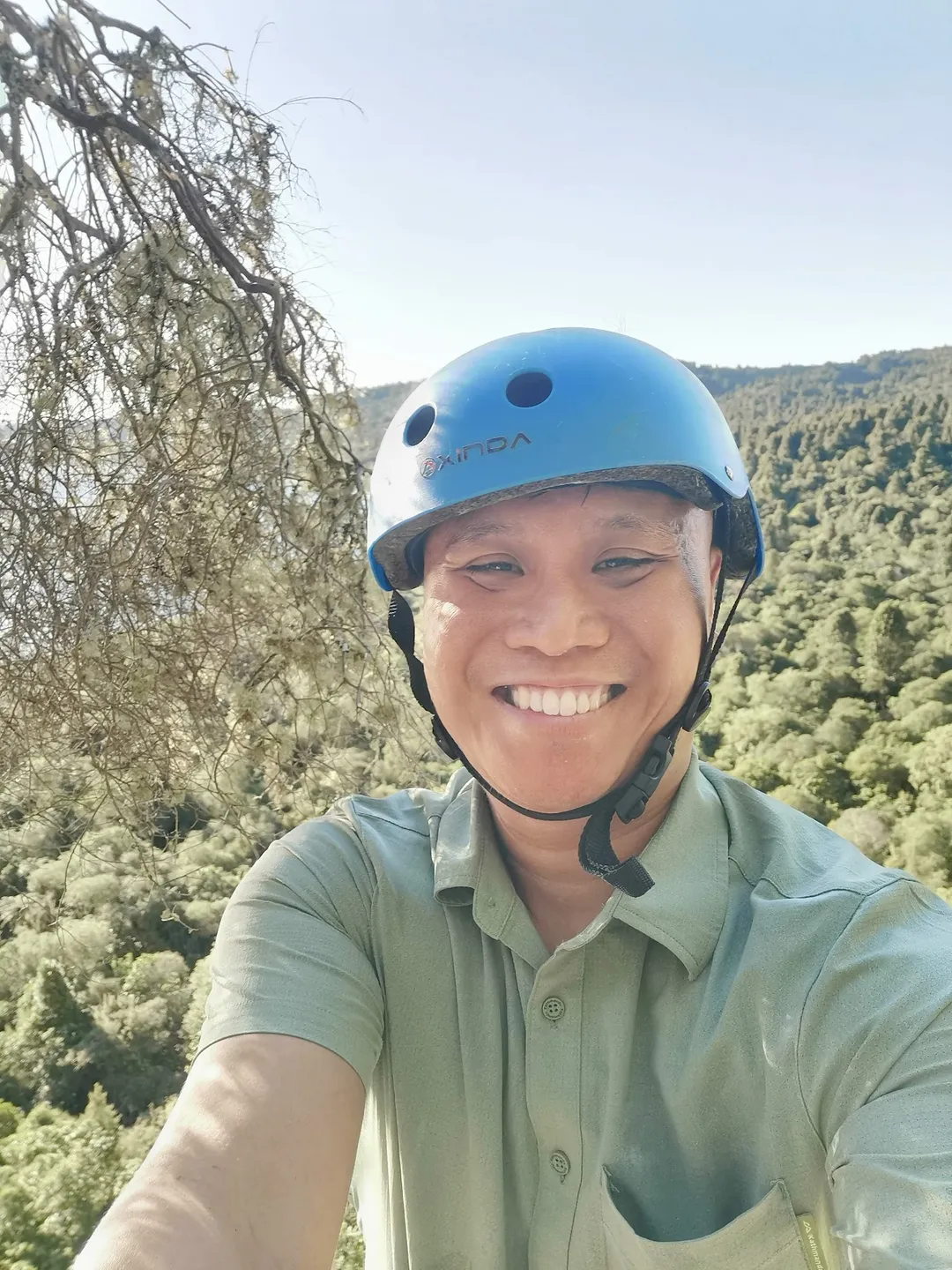 Zhi Lee in a climbing helmet on a cliff ledge above the forest at Kawakawa Bay